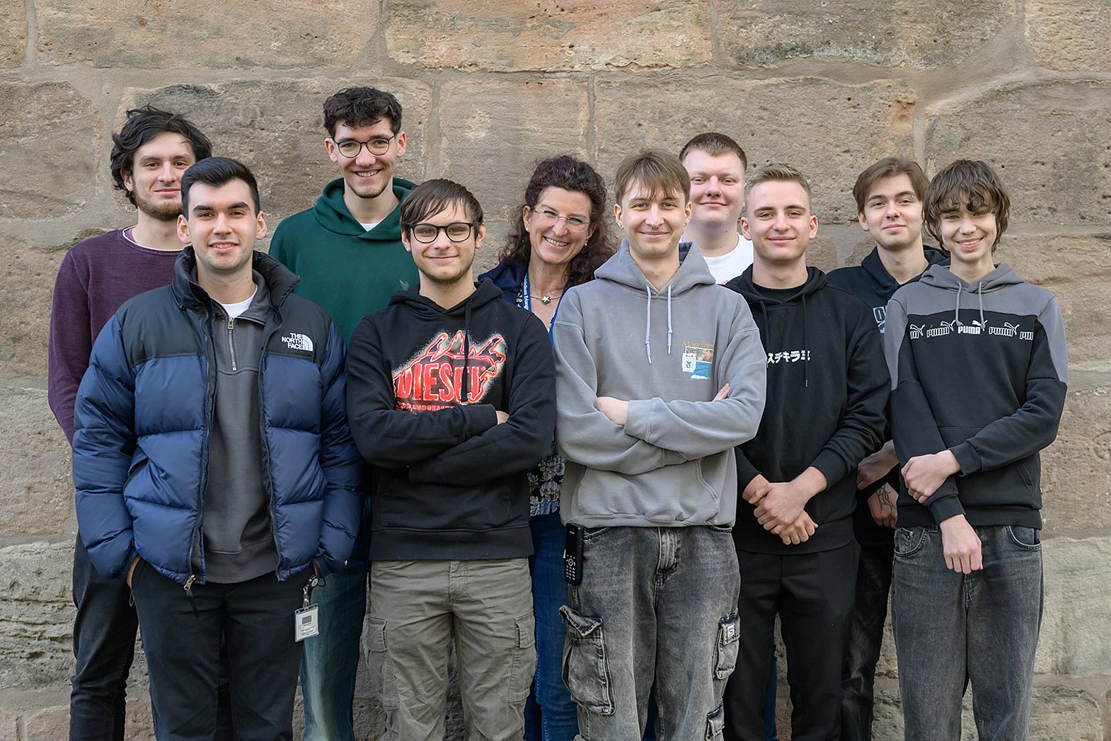 Es ist ein Gruppenfoto von neun MIK-Azubis vor einer Steinwand zu sehen, zusammen mit Ausbilderin Katharina Diesch in der Mitte