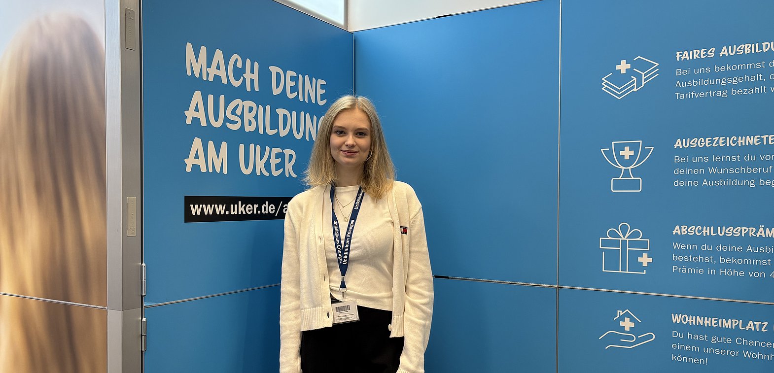 Emely Hofbauer_Messebetreuung_Vocatium Erlangen 2025 Junge Frau steht vor einer Messewand des Uniklinikums Erlangen und lächelt freundlich in die Kamera